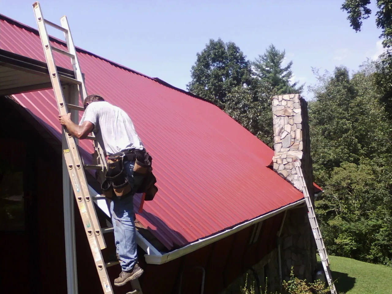 Roofer on a ladder beside a red metal roof and stone chimney — Soffit & Fascia Repair by Roofing Brothers in Cerro Gordo
